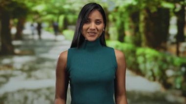 Yeşil elbiseli gülümseyen genç Latin kadın, bereketli ağaçlarla çevrili güneşli bir park ortamında neşeli bir şekilde etkileşime geçer ve canlı bir açık hava ortamı yaratır..
