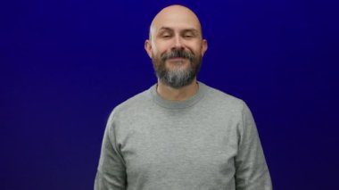 Sakallı kel adam mavi arka planda jest yapıyor gri süveter giyiyor cazibesi ve kendine güveni var..