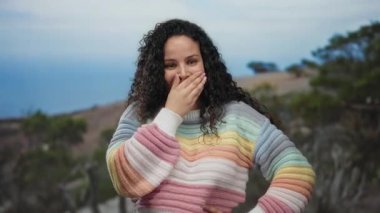 Sahilde renkli bir süveter giyen, deniz manzaralı bir kadın gündüz vakti neşeli bir hava sergiliyor..