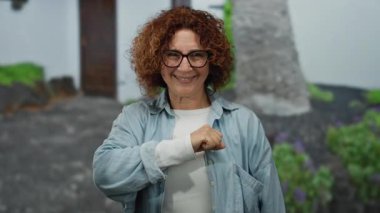 Kıvırcık saçlı İspanyol kadın şehir caddesinde kameraya yumruğunu sergiliyor. Açık hava ortamında güç ve pozitifliği somutlaştırıyor..