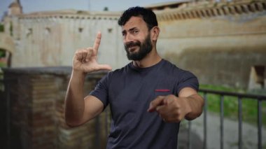 Roma 'da antik mimarinin önünde şakacı bir jest yapan sakallı İspanyol bir adam, tarihi bir taş arka plana işaret ediyor ve gülümsüyor..