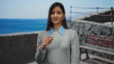 Deniz kenarında deniz kenarında gezinen bir kadın, parmak izi yok, genç Latin, gri kazak giyiyor, plaj manzarası var. Rahat bir hava yaratıyor..