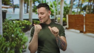 Dövmeli genç adam gülümsüyor ve yeşil arka planda ağaçlar ve yeşil bir şehir ortamında başparmak kaldırıyor, pozitiflik ve güven taşıyor..