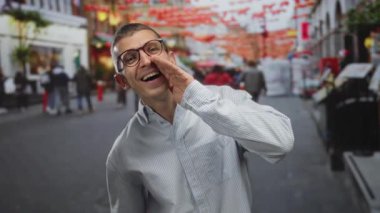 Gözlüklü, çizgili tişörtlü, gülümseyen ve arka planda kırmızı süslemeleri olan kalabalık bir sokakta konuşan bir adam gün ışığında canlı şehir manzarası yakalıyor..