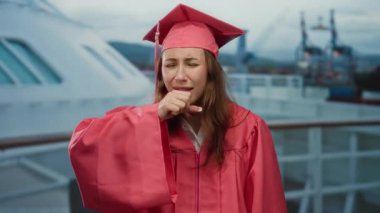 Pembe mezuniyet elbisesi giymiş bir kadın yolcu gemisinin güvertesinde öksürüyor, kendini iyi hissetmiyor, kenetlenmiş bir yolcu gemisinin arka planı ve bulutlu bir gökyüzünün altında uzak vinçlerle..