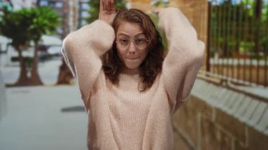 Kentsel bir sokak ortamında komik suratlar yapan, gözlüklü ve kıvırcık saçlı bir kadın sonbahar modasına uygun bir şehir ortamında neşeli bir ifade sergiliyor..