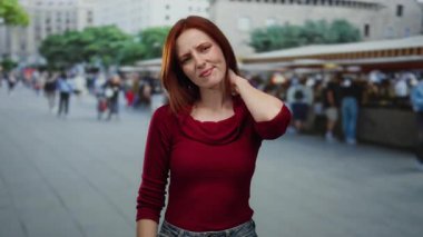 Açık pazar caddesinde boynuna dokunan kızıl saçlı, kırmızı kazaklı bir kadın yoğun şehir sahnesinde bulanık arka plan yayaları arasında acı içinde beliriyor.