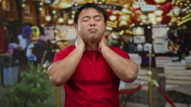 Kırmızı gömlekli genç adam, arka planda parlak ışıklarla kalabalık bir caddede boyun ağrısı çekiyor ve kentsel rahatsızlığı ve yorgunluğu aktarıyor..