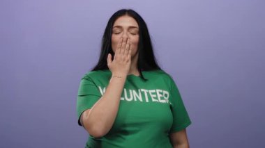 Genç bayan gönüllü kameraya bir öpücük yolluyor. Yeşil bir gömlek giyip mor bir arka plana yaslanıyor.
