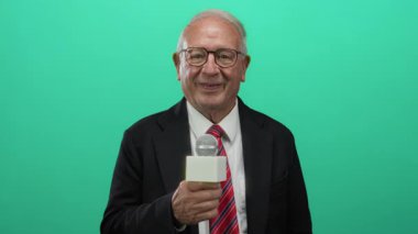 Takım elbiseli kıdemli bir adam yeşil bir arka plan duvarına mikrofon tutarak kameraya dostça ve profesyonel bir şekilde el sallıyor..
