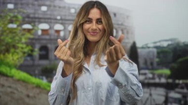 Sarışın genç bir kadın, iki eliyle güneş ışığı altındaki kolezyumun önünde kalp hareketi yapar..