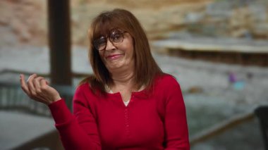 Kırmızı tişörtlü, kıdemli İspanyol kadın neşeyle sahilde poz veriyor. Gülümsüyor, sakin sahil atmosferini kucaklıyor..