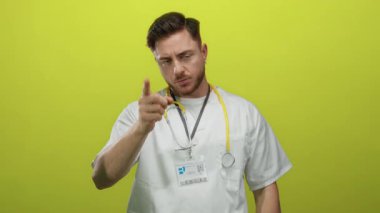 Sakallı, beyaz, genç bir doktor. Parmak ucunda, izole edilmiş sarı arka plana karşı steteskop takıyor. Ciddi bir ifade ve profesyonel bir varlık sergiliyor..