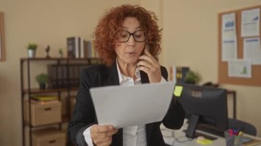 Modern ofis ortamında telefon başında doküman okuyan bir kadın, etrafındaki raflar, bitkiler ve bilgisayarlarla kapalı bir çalışma ortamı sergiliyor..