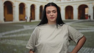 Tarihi bir üniversite avlusunda kendine güvenen bir kadın, sıradan bir bej gömlek giyiyor, kemerler ve kaldırım taşları arasında büyük boy güzellik ve dingin bir güven simgeliyor.