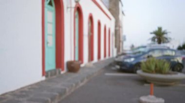 Lanzarote 'deki bulanık sokak manzarası, kırmızı kemerler ve turkuaz kapılar, arabalar, bitkiler ve palmiye ağaçları olan renkli bir binaya odaklanan İspanya.