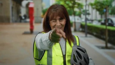 Güvenli bir yelek giyen kadın, güneşli bir günde şehir manzarası ve ağaçlarla çevrili, göz kırpan ve işaret eden bir şehir caddesinde bisiklet kaskı tutuyor..