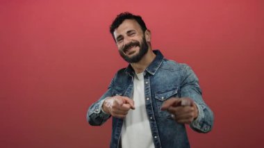 Sakallı İspanyol bir adam gülümsüyor ve kırmızı arka planda kot ceket giyiyor..