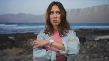 Gündelik giysiler giyen bir kadın kayalık bir sahilde dikiliyor. Arka planda dalgalar ve dağlar, berrak bir gökyüzünün altında huzurlu bir deniz kenarı anını yakalıyor..