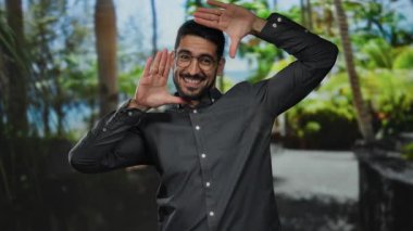 İspanyol genç adam elleriyle jest yapıyor, yemyeşil bir açık hava parkında gülümsüyor..