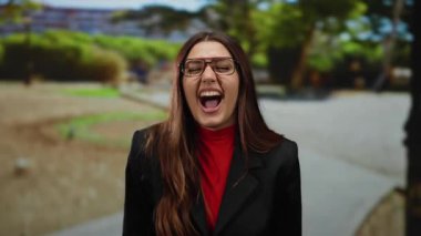 Parktaki İspanyol kadın gülüyor ve gülümsüyor, gözlükler takıyor, uzun saçlı ve güneşli bir günde siyah ceketin altında kırmızı bir gömlek giyiyor..
