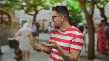 Şehir sokağında gözlüklü genç bir adam. Arka planda bulanık insanlar var. Dövmeleri var, çizgili bir gömlek giyiyor..