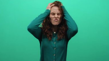 İzole edilmiş yeşil arka plan duvarına karşı elleri başındaki hayal kırıklığını ifade eden kadın, tarafsız bir bağlamda stres ve duygu taşıyor..