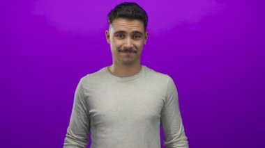 Genç İspanyol adam iki parmağıyla aşağıya doğru işaret ediyor. Kalın mor arka plan duvarına karşı. Açık gri gömlek giyiyor.