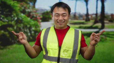 Yeşil bir parkta, güvenlik yeleği giymiş, gülümseyen ve iki eliyle işaret eden genç adam çeşitli yönleri ve jestleri gösteriyordu..