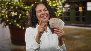 Şehir sokaklarında elinde Kolombiya pezosu tutan orta yaşlı bir kadın açık havada neşeli ışıklar altında gülümsüyor. Mutluluk ve finansal başarıyı yansıtıyor..