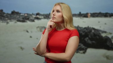 Kayalık bir sahilde arka planda okyanus manzarası olan bir kadın, kırmızı bir gömlek giyiyor, düşünceli bir ifade sergiliyor ve sakin bir deniz manzarası ile çevrili..