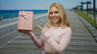 Kumsalda elinde hediyeyle gülümseyen bir kadın okyanus manzaralı pembe bir hediye sunuyor. Mutluluğu ve deniz kenarındaki mutluluğu yansıtıyor..