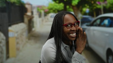 Kırmızı gözlüklü ve örgülü gülümseyen bir kadın, arka planda arabaları olan bir şehir sokağında neşeli bir şekilde el kol hareketi yapar, açık havada neşeli ve çekici bir anı yakalar..