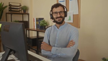 Kulaklık takan İspanyol adam modern ofiste bilgisayar masasında kollarını kavuşturarak gülümsüyor. Kitaplık ve bitkilerle birlikte profesyonel ve misafirperver bir ortam yaratıyor..