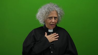 Siyah cüppeli kadın göğsünün üzerinde yeşil bir duvara karşı duruyor. Duygusal ifadesi samimiyet ve bağlılık gösteriyor..