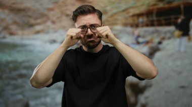 Deniz kenarında sakallı ve gözlüklü genç adam yaz aylarında bulanık arkaplanlı sahilde güneşli gökyüzünün altında üzgün görünüyor..