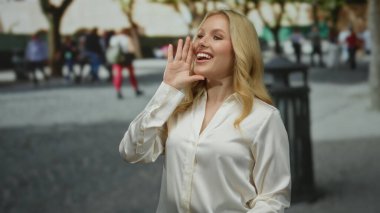 Yoğun bir şehir caddesinde beyaz gömlek giyen genç ve sarışın bir kadın ağaçlar ve arka planda insanlarla dolu canlı bir şehir ortamında neşe içinde bağırıyor..