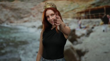 Taçlı bir kadın, gün ışığında sahilin doğal bir sahile karşı duran neşeli bir hareketle sahilde duruyor..