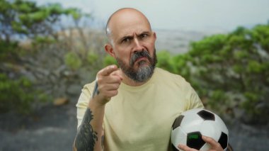 Sakallı kel bir adam parkta futbol topu tutarak tehdit edici hareketlerde bulunuyor. Dışarıdaki duygulara ve dövmelere vurgu yapıyor..