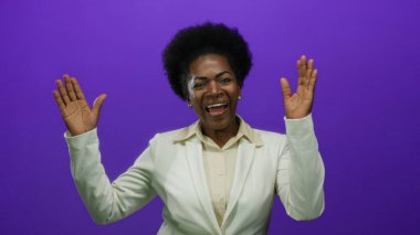 Beyaz takım elbiseli bir kadın canlı mor bir arka plana gülümsüyor, mutluluk ve profesyonellik sergiliyor, ellerini canlı bir şekilde kaldırıyor..