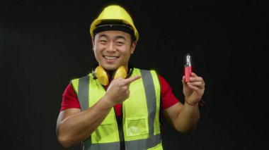 Kasklı ve güvenlik yeleği olan gülümseyen inşaatçı elinde siyah arka plan aydınlatma cihazıyla ürünü vurguluyor.