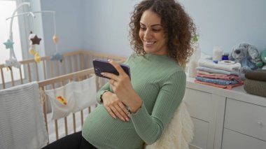 Hamile bir kadın gülümsüyor, rahat bir yatak odasında oturuyor, telefonuyla meşgul, yumuşak ışık, beşik ve bebek malzemeleriyle çevrili, huzurlu bir ev ortamı yaratıyor..