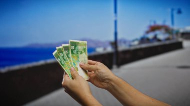El ele tutuşan İsrail şekeli banknotlar deniz kenarında, bir iş ya da yatırım teması yakalamak bir kıyı arkaplanı karşı.
