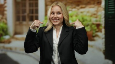 Akdeniz ev ortamında gülümseyen, kendini gösteren, siyah takım elbiseli, taş arka planı ve yeşil bitkileri olan, profesyonel ama sade bir hava yakalayan bir kadın..