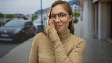 Sarışın genç bir kadın kenar mahallede yanağa dokunuyor, dedikodu gizliliği fısıldıyor..