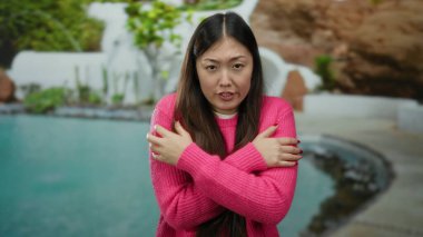 Pembe kazaklı kadın havuz kenarında poz veriyor. Soğuk, gözleri kapalı ve kolları çaprazlanmış bir şekilde sakin bir otelde huzurlu Asyalı güzelliğini sergiliyor.