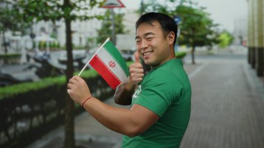 Başparmaklarıyla gülümseyen genç adam İran bayrağını şehir caddesinde tutuyor.