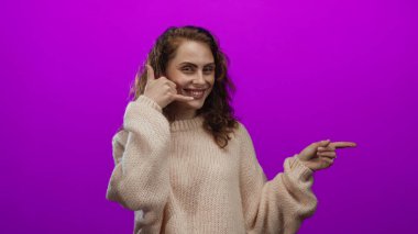 Bir telefon konuşması yaparken gülümseyen ve parlak pembe bir arkaplanı işaret eden, neşeli ve cana yakın bir ifadeyle rahat bir kazak giyen bir kadın..