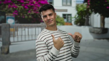 Gülümseyen ve el kol hareketi yapan genç bir adam canlı bir şehir caddesinde çiçekli bir arka planda gün ortasında çekilen çekimlerde neşe ve pozitiflik saçan.