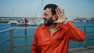 Turuncu tulumlu ve sakallı bir adam limanda duruyor. Gemiler ve teknelerle dolu ve deniz zeminine karşı el ele hareketlerini dinlerken..